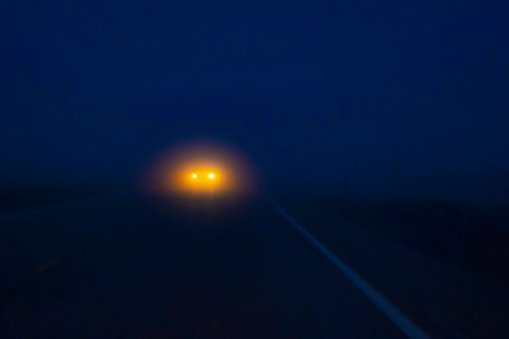 Car headlights glow yellow-orange in the middle distance at night, nothing else visible save for a diagonal border of the road’s edge 